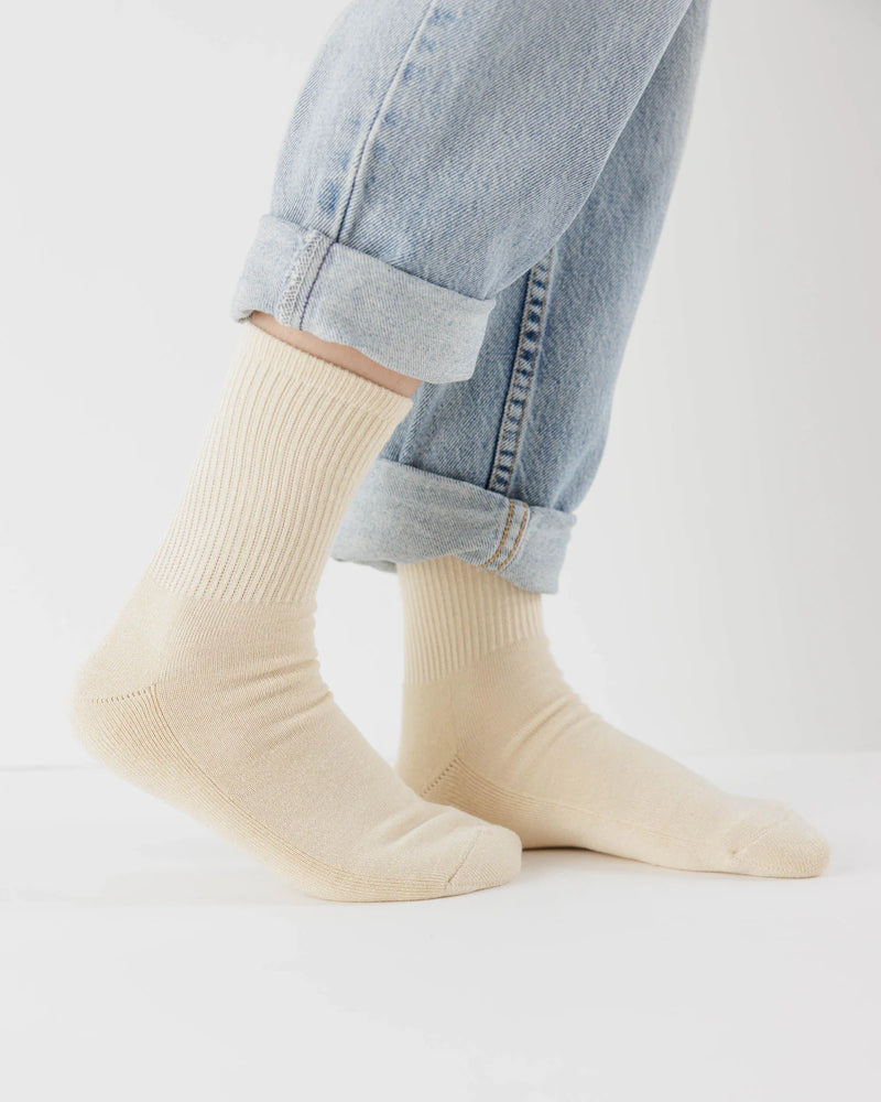 Ribbed Socks Ecru an den Füßen einer Person, die hochgekrempelte Jeans trägt. Diese beigen, gerippten Socken bieten Komfort dank weicher, dehnbarer Bambus-Rayon-Mischung.