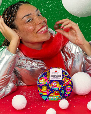 9 Milchschokoladekugeln mit Haselnusspralinenfüllung in Weihnachtsdose, präsentiert von einer lächelnden Frau, die eine Praline hält, umgeben von dekorativen Schnee-Requisiten.