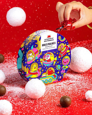 Runde Weihnachtsdose mit 9 Milchschokoladekugeln, Haselnusspralinenfüllung. Weihnachtsillustration mit fliegenden Untertassen auf rotem Untergrund, umgeben von Schokokugeln und Schneebällen. Hand greift Praline.