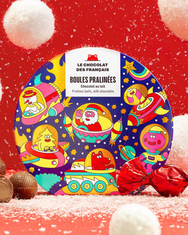 Weihnachtsdose mit 9 Milchschokoladekugeln und Haselnusspralinenfüllung, illustriert mit Cartoon-Figuren. Einige Pralinen sind ausgepackt, eine in roter Folie eingewickelt.