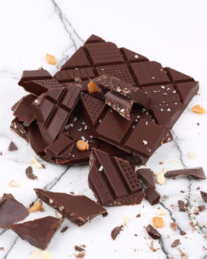 Triomphe Dunkle Schokolade mit Mandeln und gesalzenem Karamell 70g: Zerbrochene Schokoladenstücke mit Karamell, Mandeln und Salz auf Steinoberfläche.