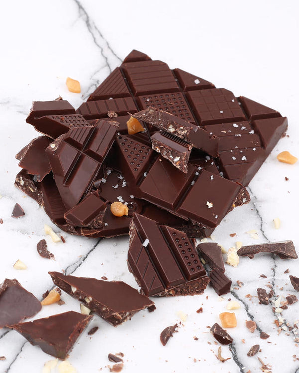 Triomphe Dunkle Schokolade mit Mandeln und gesalzenem Karamell 70g: Zerbrochene Schokoladenstücke mit Karamell, Mandeln und Salz auf Steinoberfläche.