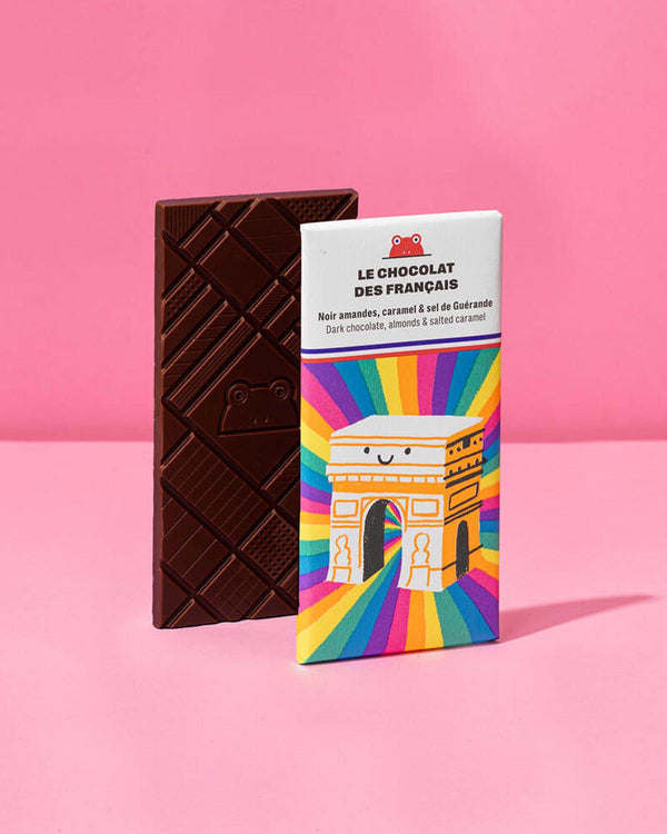 Triomphe Dunkle Schokolade mit Mandeln und gesalzenem Karamell 70g, zeigt geometrisches Froschmuster. Verpackung illustriert mit lachendem Arc de Triomphe von Carolina Buzio.