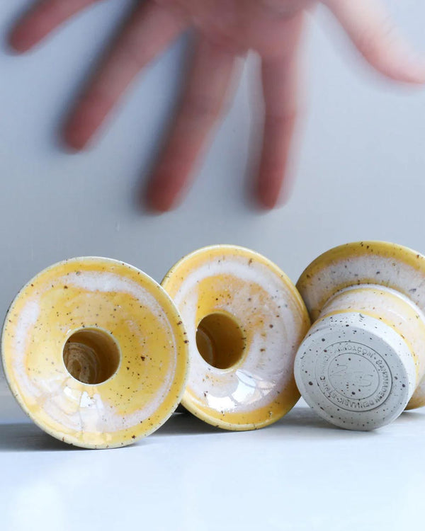 Hono Cornflower Cream Kerzenständer aus Keramik, handgefertigt von Studio Arhoj, mit speckelnder gelb-weißer Glasur, auf einem Tisch liegend, Hand greift danach.