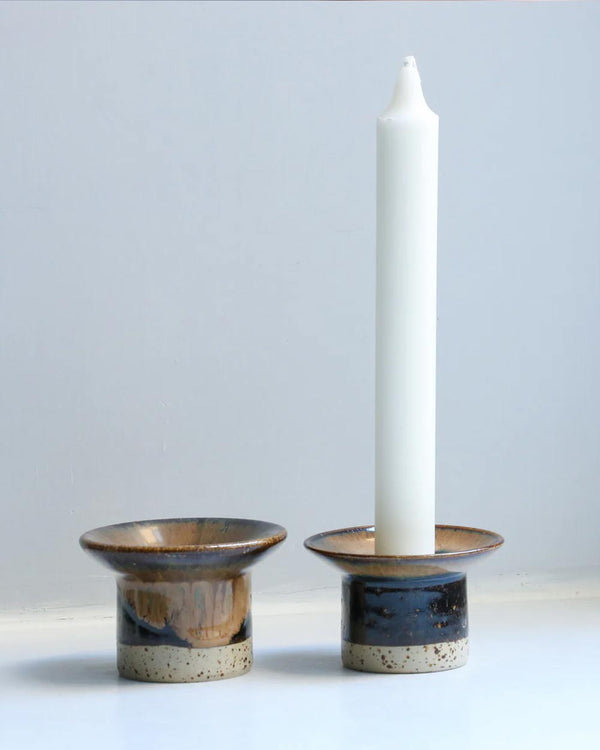 Zwei handgefertigte Hono Kerzenständer von Studio Arhoj, einer mit weißer Kerze, auf heller Oberfläche. Schlichter, funktionaler Stil mit einzigartigem Glasur-Ringeffekt.