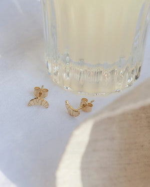 Croissant Ohrstecker Gold aus vergoldetem Sterlingsilber, elegant auf hellem Stoff präsentiert, mit einem klaren, geriffelten Glasgefäß im Hintergrund.