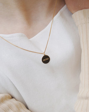 MOM Kette Gold: Feine, vergoldete Halskette mit rundem MOM-Anhänger, getragen zu einem weißen Oberteil. Entworfen in Berlin, handgefertigt in Italien.