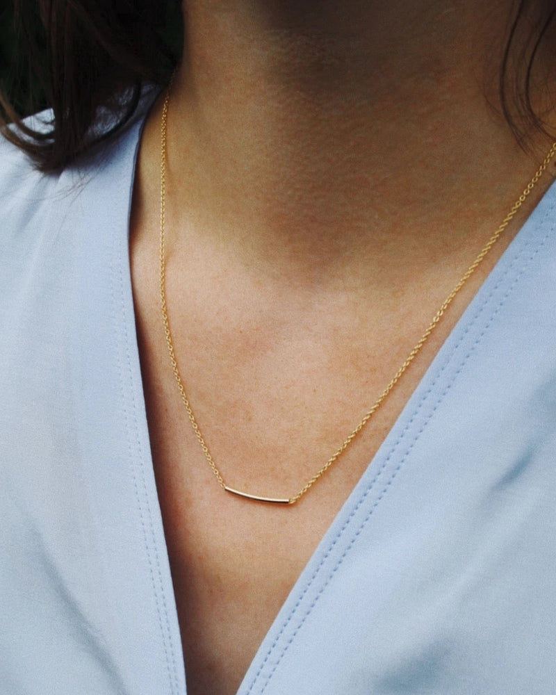 Nahaufnahme einer zarten Pipe Kette Gold mit horizontalem Stab-Anhänger aus 24 Karat vergoldetem Sterlingsilber, getragen über einem hellblauen V-Ausschnitt.