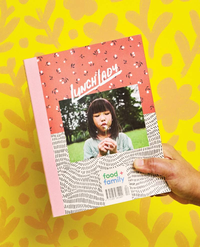 Person hält Lunch Lady No. 28 Magazin mit Kind, das Pusteblume betrachtet. Themen: Familie, Ernährung. Erhältlich bei Human Empire Shop.