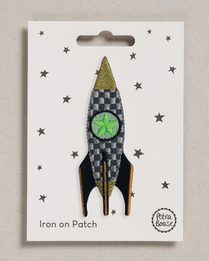 Rakete Aufnäher (zum Aufbügeln): Gestickter Patch in Raketenform mit grünem Fenster, schwarzem-goldenem Finnen, auf sternenbedrucktem Karton, Petra Boase Logo unten rechts.