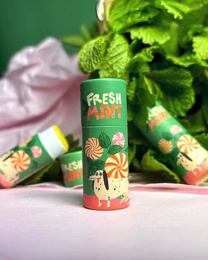 Lip Balm Fresh Mint: Minzgrüne Röhrchen mit Minzmotiv und Dackel, umgeben von frischen Minzblättern. Handgefertigt in Schweden aus biologischen Zutaten.