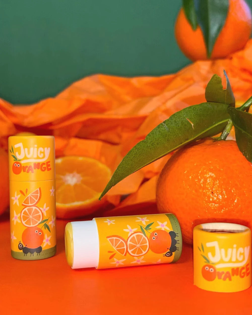 Drei Juicy Orange Lippenbalsam-Stifte mit Cartoon-Orangen, umgeben von einer geschälten und einer ganzen Orange, auf einem orangefarbenen Hintergrund arrangiert.