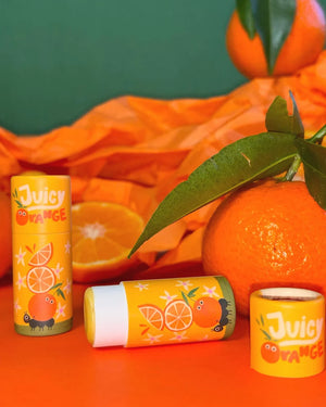 Drei Juicy Orange Lippenbalsam-Stifte mit Cartoon-Orangen, umgeben von einer geschälten und einer ganzen Orange, auf einem orangefarbenen Hintergrund arrangiert.