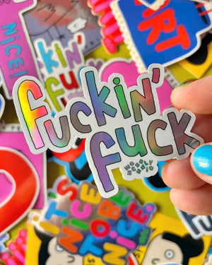 Hand hält Fuckin Fuck Holo-Sticker von Marie Chanteur aus holografischem Vinyl, umgeben von weiteren verspielten Stickern auf einer Oberfläche. Größe: 7,4 x 6,2 cm.