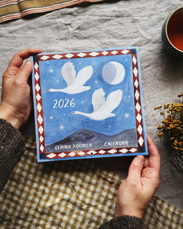 Gemma Koomen 2026 Kalender mit Nachthimmel-Illustration, gehalten von Händen, auf einem Tisch mit Tee und getrockneten Blumen, platzsparend und umweltfreundlich gedruckt.