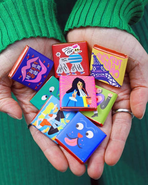 Le Chocolat des Francais Minis (8x5g) in bunten Folien mit Illustrationen, gehalten in Händen mit grünem Pullover; köstliche Schokoladenmischung aus Frankreich.