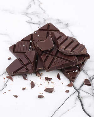 Notre Dame Extra dunkle Schokolade mit Salz aus der Camargue, unregelmäßig gebrochene Stücke auf Marmoroberfläche, mit feinen Salzflocken verziert. 70g Tafelschokolade.