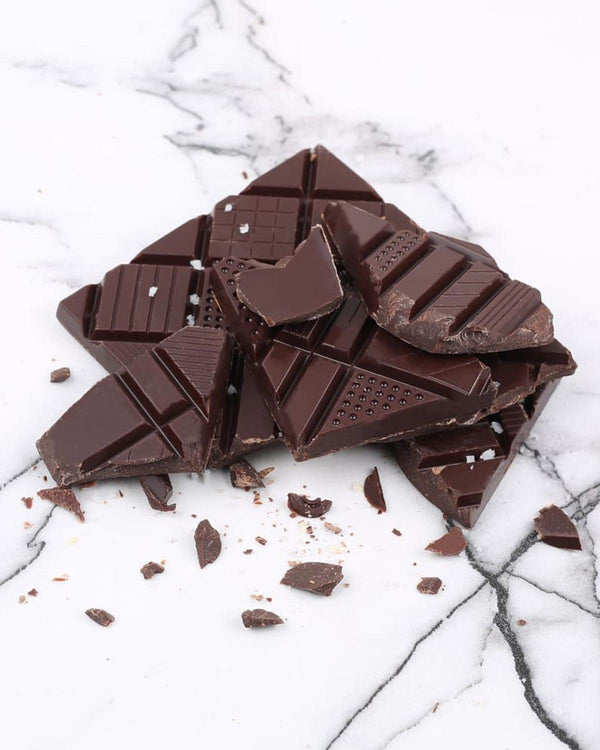 Notre Dame Extra dunkle Schokolade mit Salz aus der Camargue, unregelmäßig gebrochene Stücke auf Marmoroberfläche, mit feinen Salzflocken verziert. 70g Tafelschokolade.