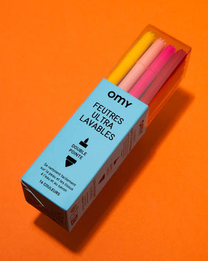 OMY - 16 Ultra Washable Markers in geöffneter Karton-Plastikbox, doppelseitige Filzstifte, leicht abwaschbar, auf orangefarbener Fläche, ideal für kreatives Gestalten.