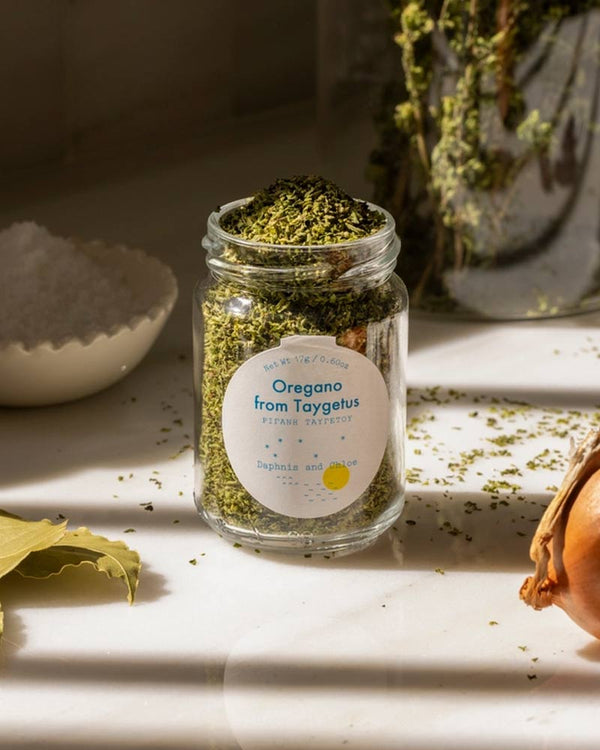 Daphnis and Chloe Oregano aus Taygetus im Glas, umgeben von grobem Salz, Lorbeerblättern und Zwiebelstück, ideal für mediterrane Gerichte und vielseitige Küchenanwendungen.