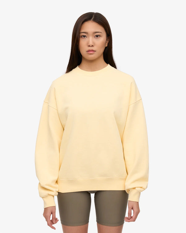Junge Frau trägt ein Oversized Sweatshirt in Soft Yellow, kombiniert mit olivgrünen Bike-Shorts. Lässiger Look von Colorful Standard, ideal für den Alltag.