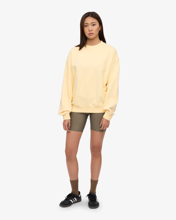 Frau trägt ein Organic Oversized Sweatshirt Soft Yellow, kombiniert mit olivgrünen Shorts, karamellfarbenen Socken und schwarzen Sneakers. Entspannter Look von Human Empire Shop.
