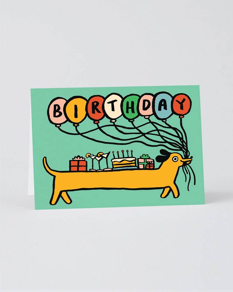 Geburtstagskarte Birthday Sausage Dog Klappkarte zeigt einen Dackel mit Geschenken und Torte auf dem Rücken, umgeben von BIRTHDAY-Ballons, illustriert von Alice Bowsher.