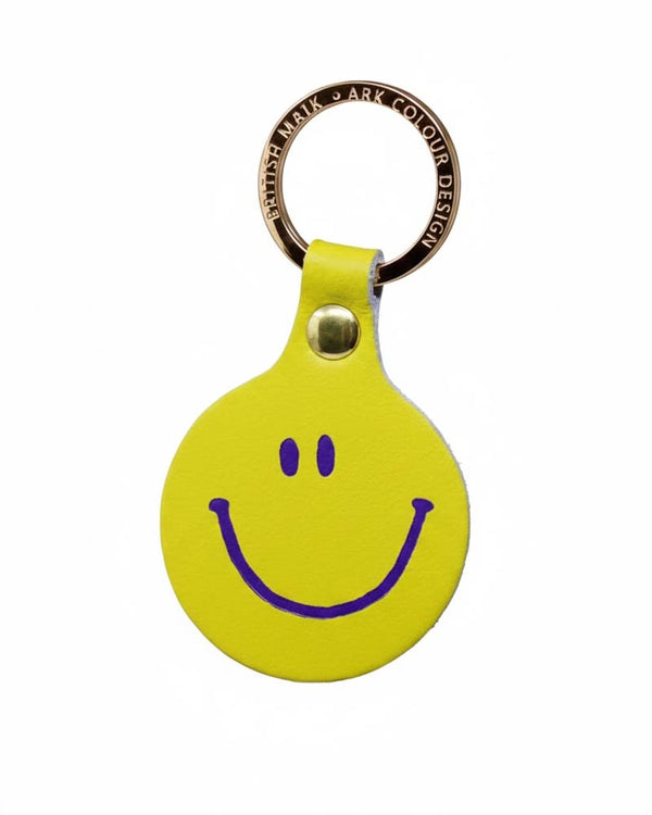Feeling Lush Smiley Schlüsselanhänger Acid Green: Runder, gelber Lederanhänger mit lila Smiley-Gesicht, befestigt an einem gravierten, messingfarbenen Schlüsselring. Hergestellt in Schottland.