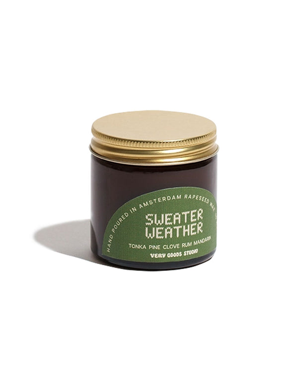 Kleine Sweater Weather Duftkerze 60ml im bernsteinfarbenen Glas mit goldfarbenem Deckel, grünes Etikett mit Duftnoten von Tonka, Kiefer, Nelke, Rum und Mandarine.